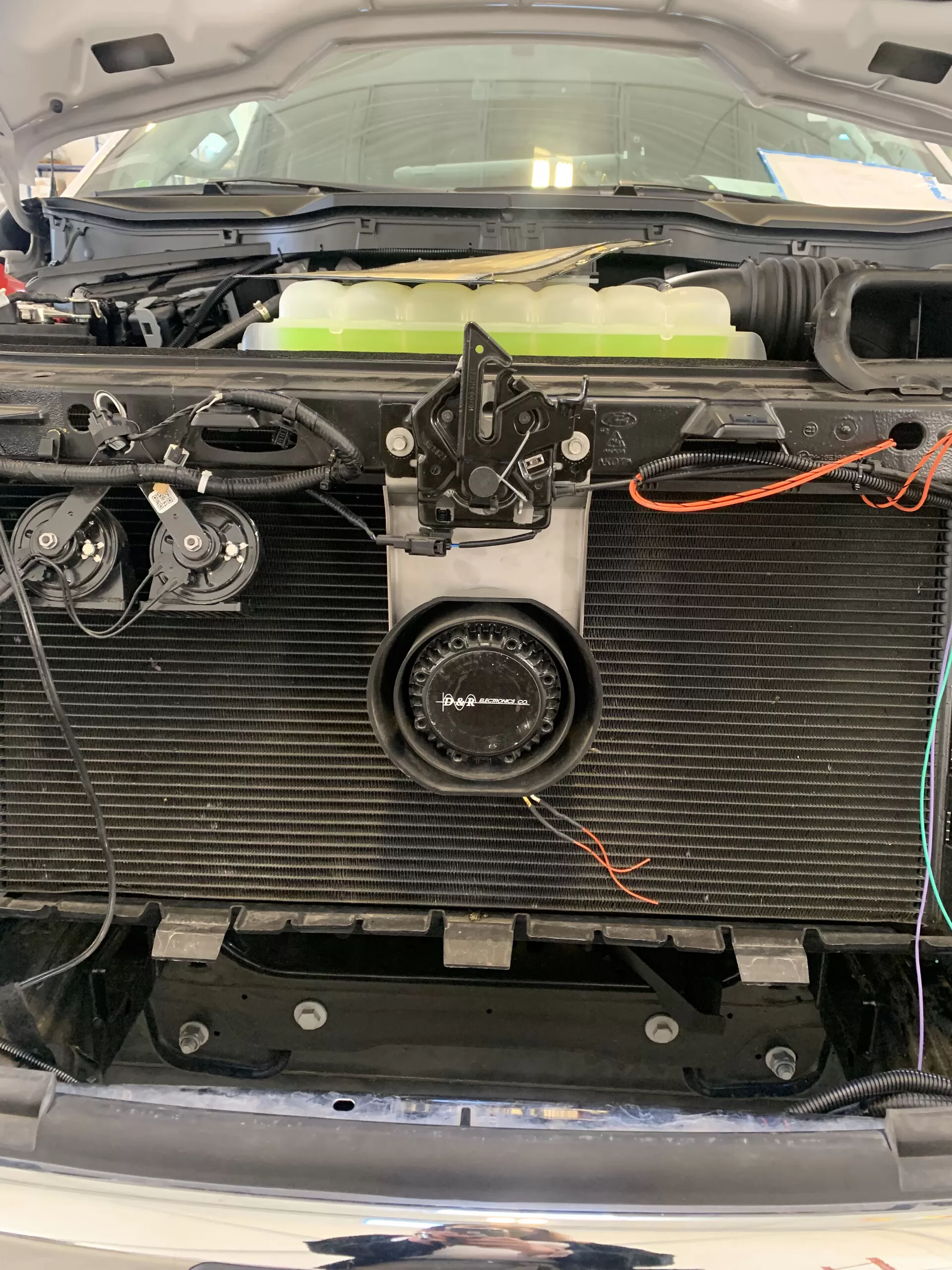 D&R-badged speaker fitted low on the radiator support, photographed under the open hood of a white vehicle